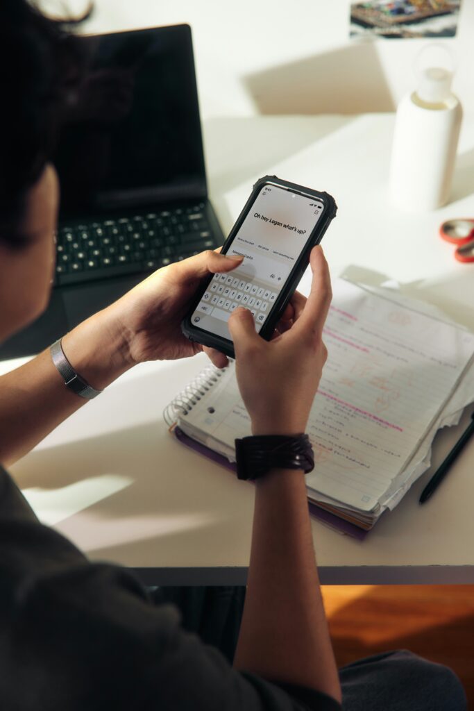 College student using AI chatbot on phone along with laptop and handwritten notes on desk.